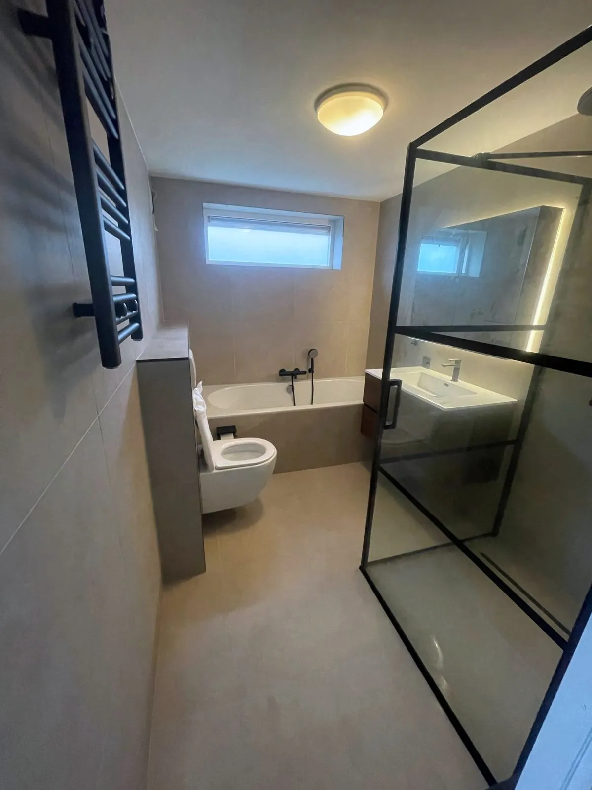 Badkamer met een ingebouwd bad, een zwevend toilet, een houten wastafelmeubel en een zwart omlijnde glazen douchewand