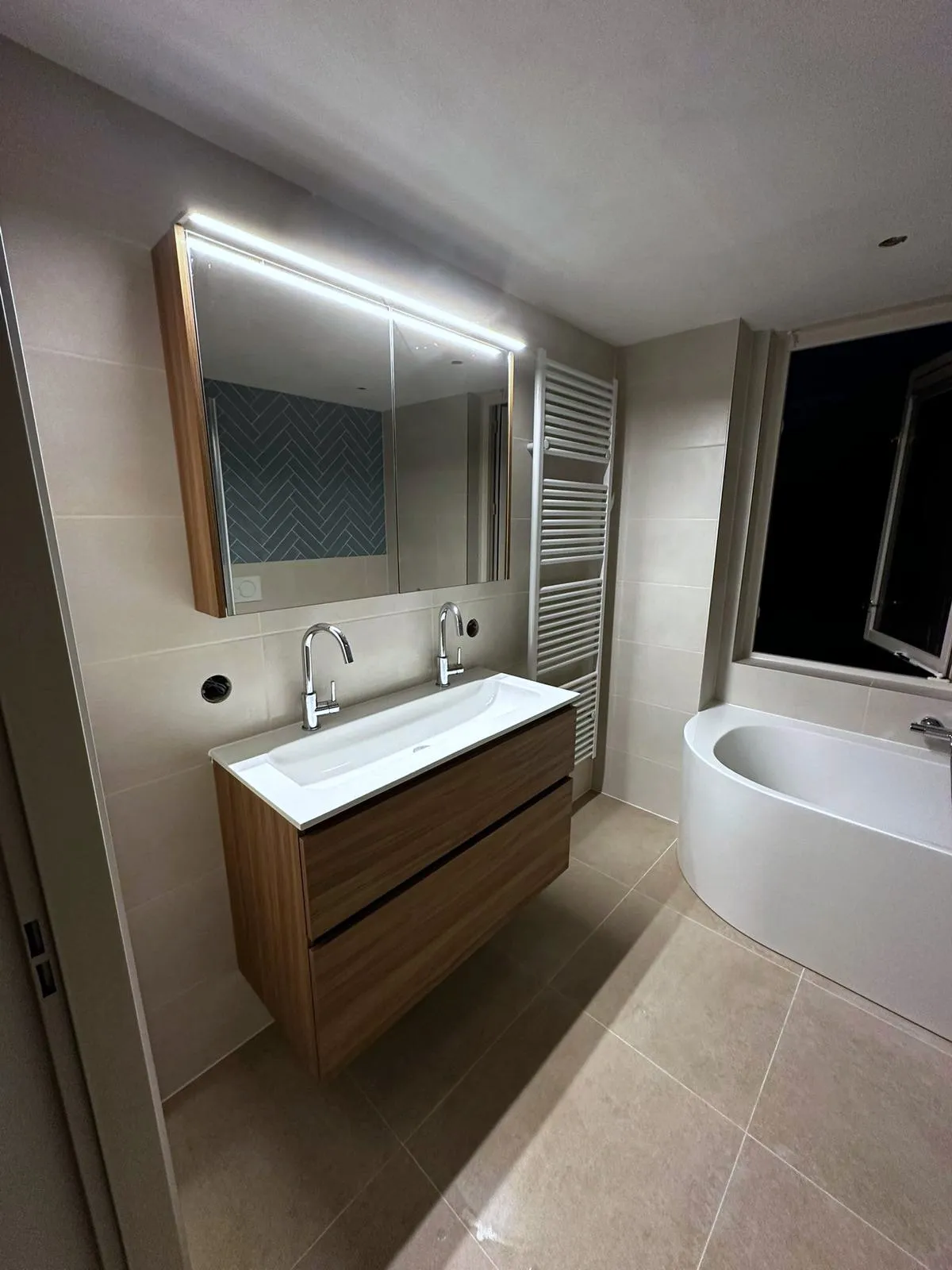 Badkamer met een dubbele wastafel, een verlichte spiegel en een bad langs de wand