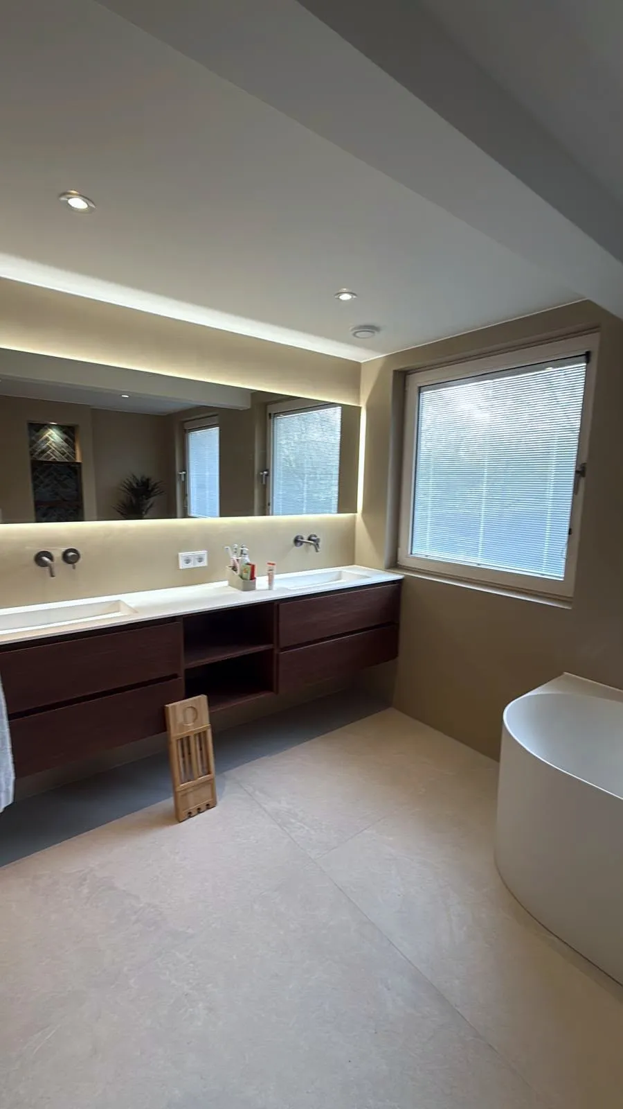 Ruime badkamer met een lang donkerhouten wastafelmeubel, een spiegelwand met verlichting, een raam en de rand van een vrijstaand bad