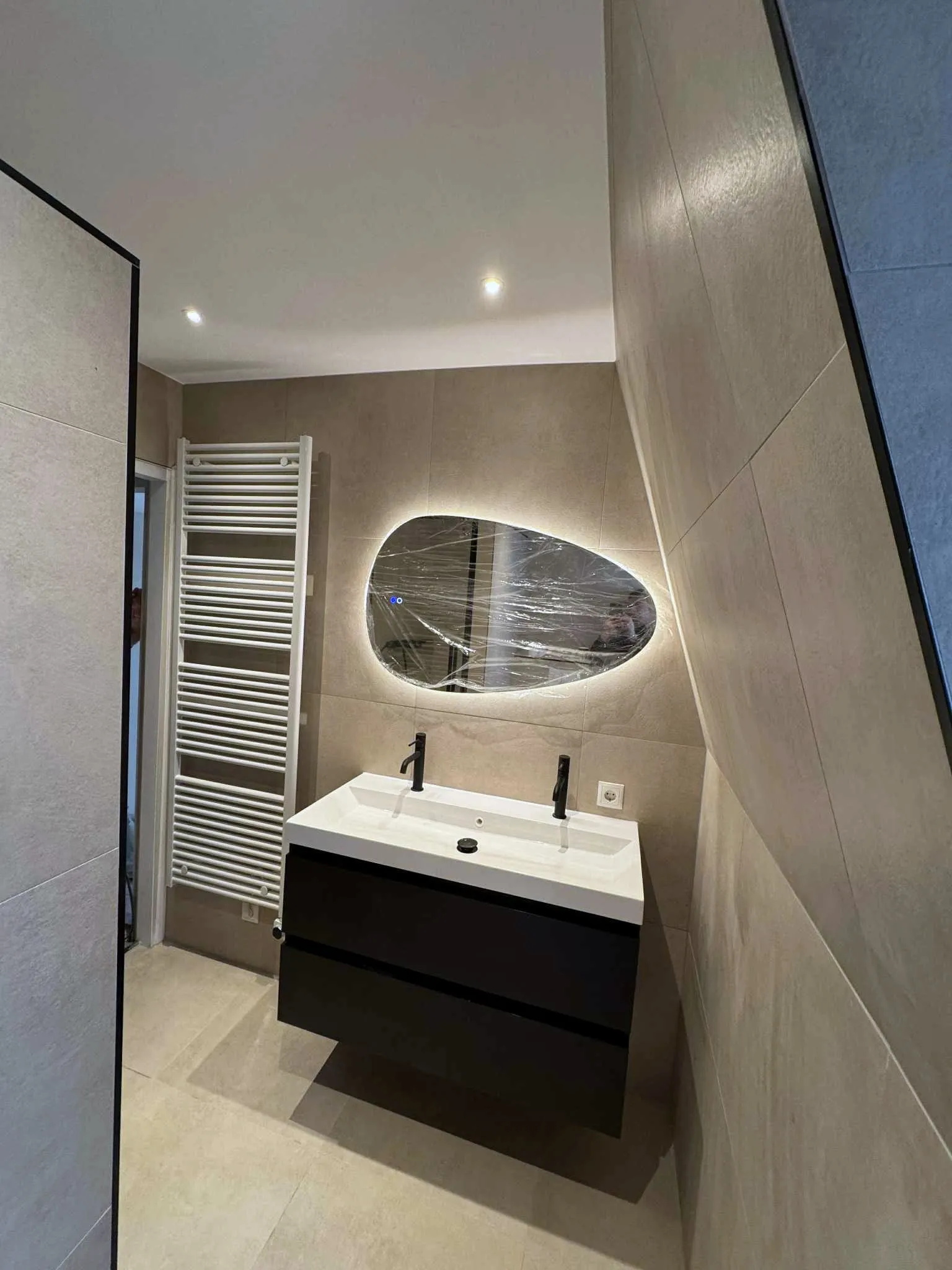 Badkamer met een ovale verlichte spiegel, een zwart wastafelmeubel en een glazen douchewand