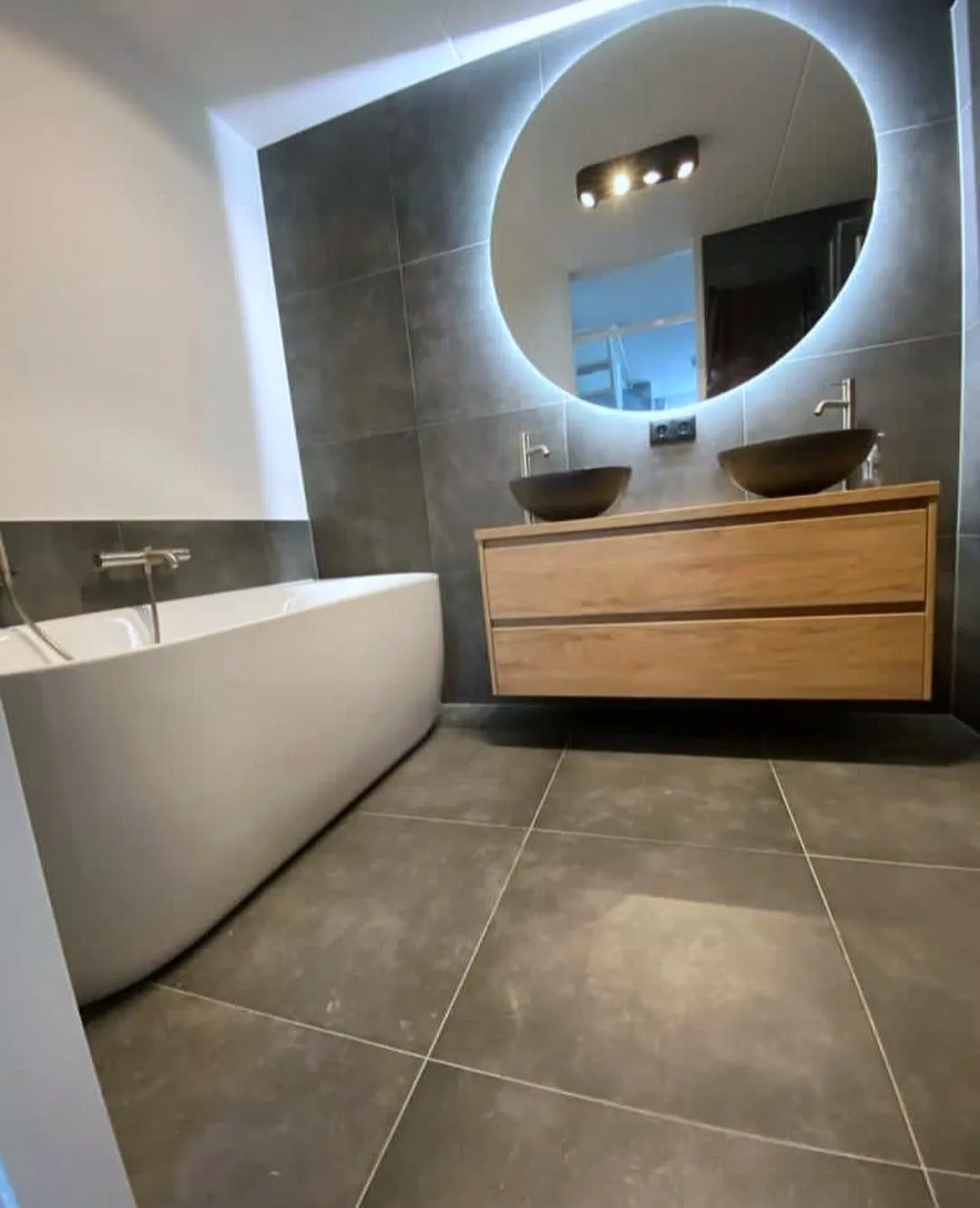 Badkamer met een grote ronde verlichte spiegel, een zwevend eiken wastafelmeubel met twee kommen en een vrijstaand bad