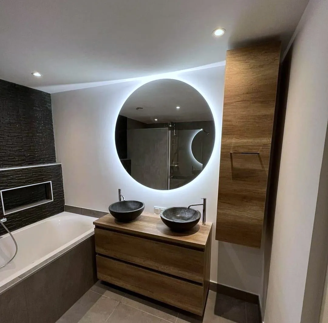 Badkamer met een ronde verlichte spiegel, een houten meubel met twee donkere steenkommen, een ingebouwd bad en een donkere mozaïekwand