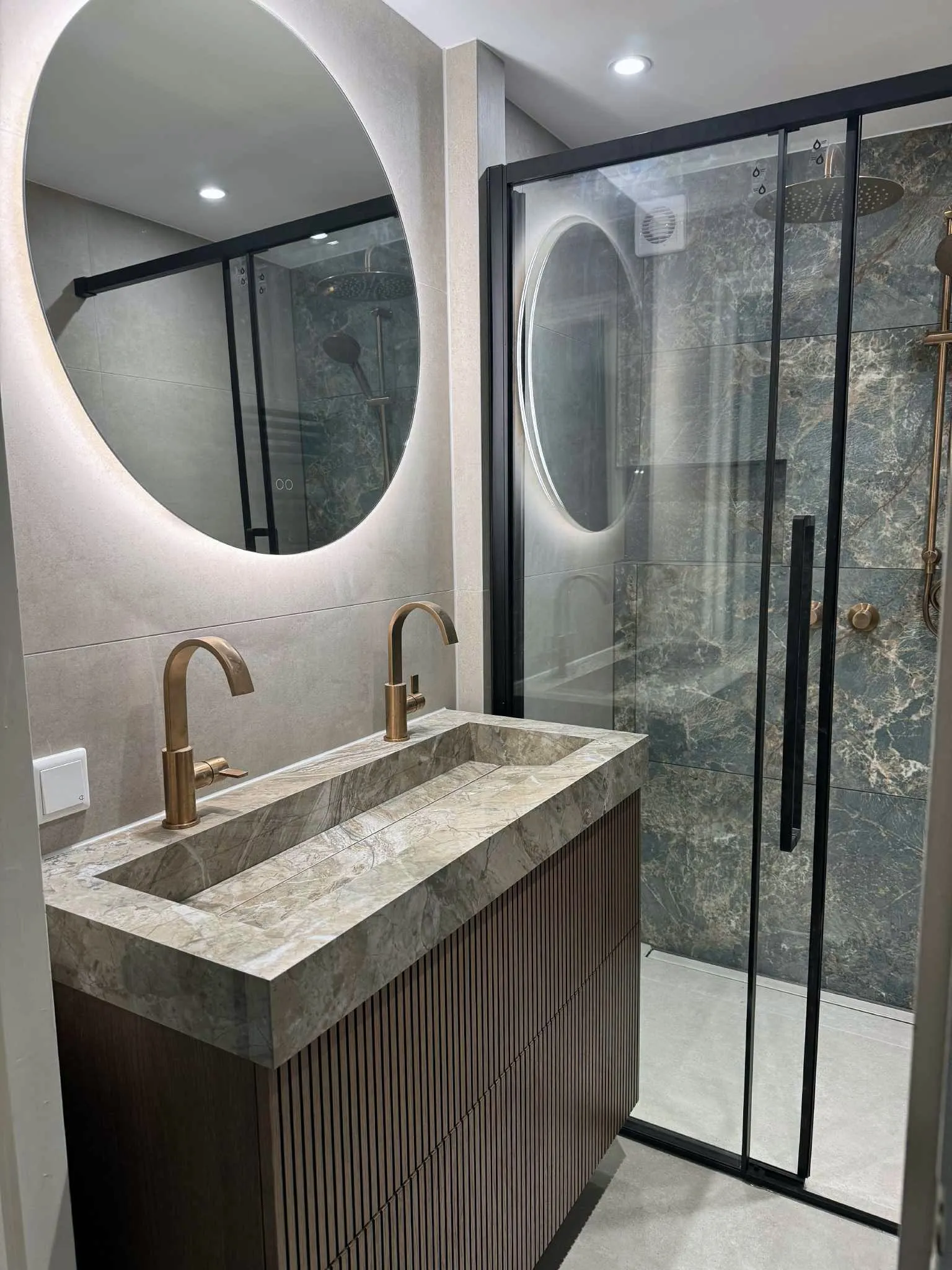 Wastafel van natuursteen met een messing kraan, ovale spiegels en een douche met zwart omlijnde glaswand
