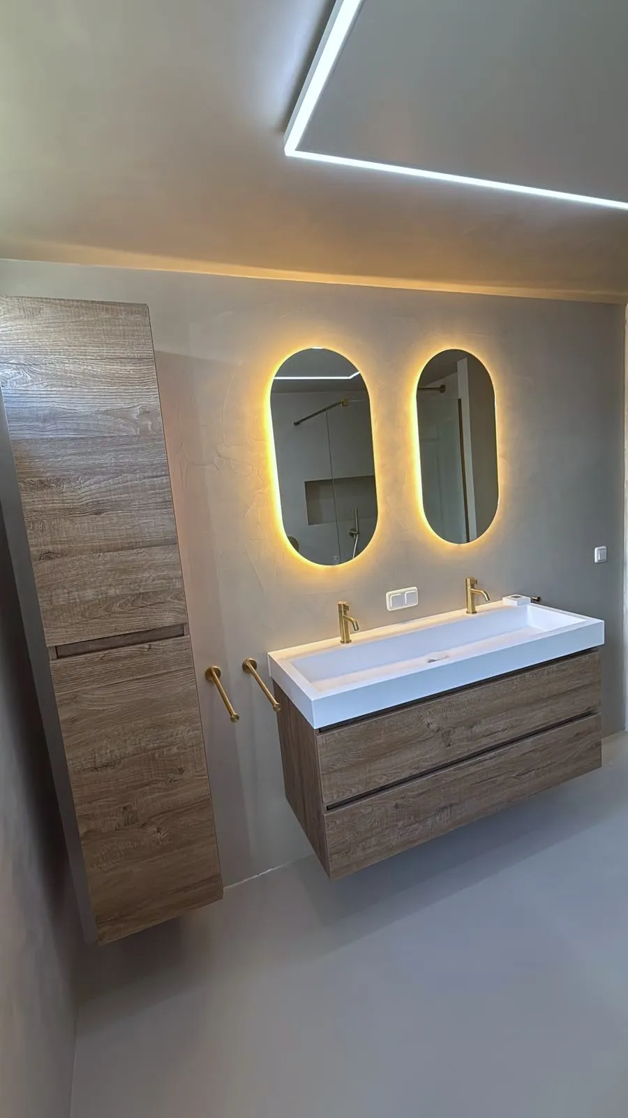 Wastafelmeubel van hout met een brede witte wasbak, twee gouden kranen en twee ovale spiegels met verlichting erachter