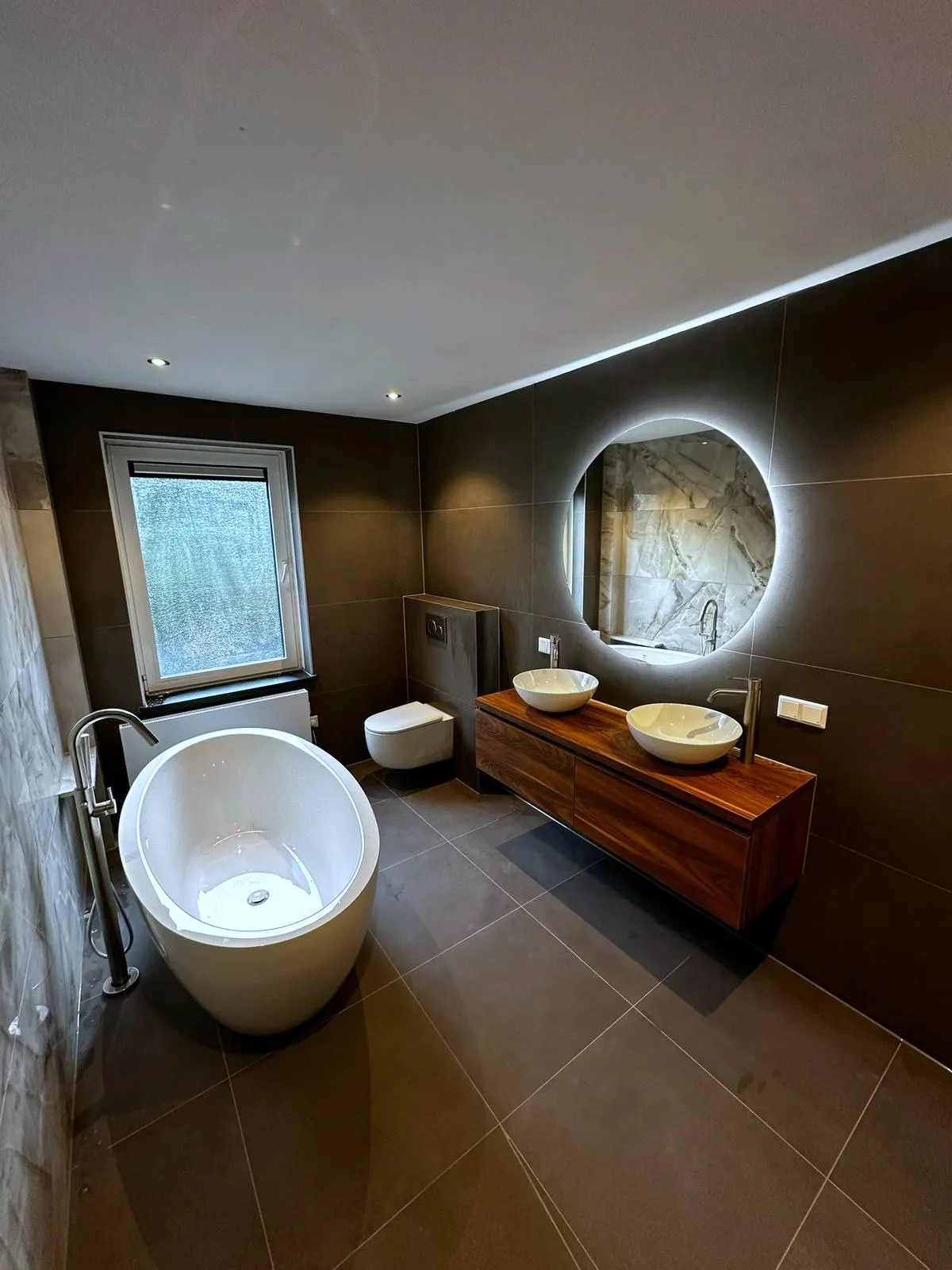 Badkamer met een vrijstaand bad, een ronde spiegel en een houten wastafelmeubel met twee kommen